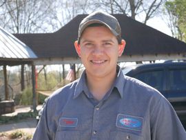 Man smiling, wearing a work shirt and baseball cap, standing outside a wooden structure on a sunny day.