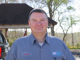 Man in blue striped work shirt smiles outdoors near a gazebo.