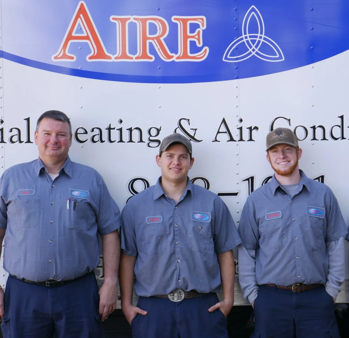Three HVAC technicians in blue shirts pose in front of their service truck. The truck displays 