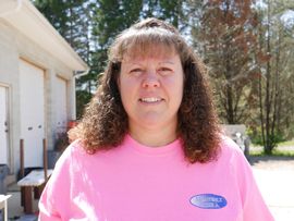 Woman with curly brown hair and pink shirt smiles in front of a building with garage doors.