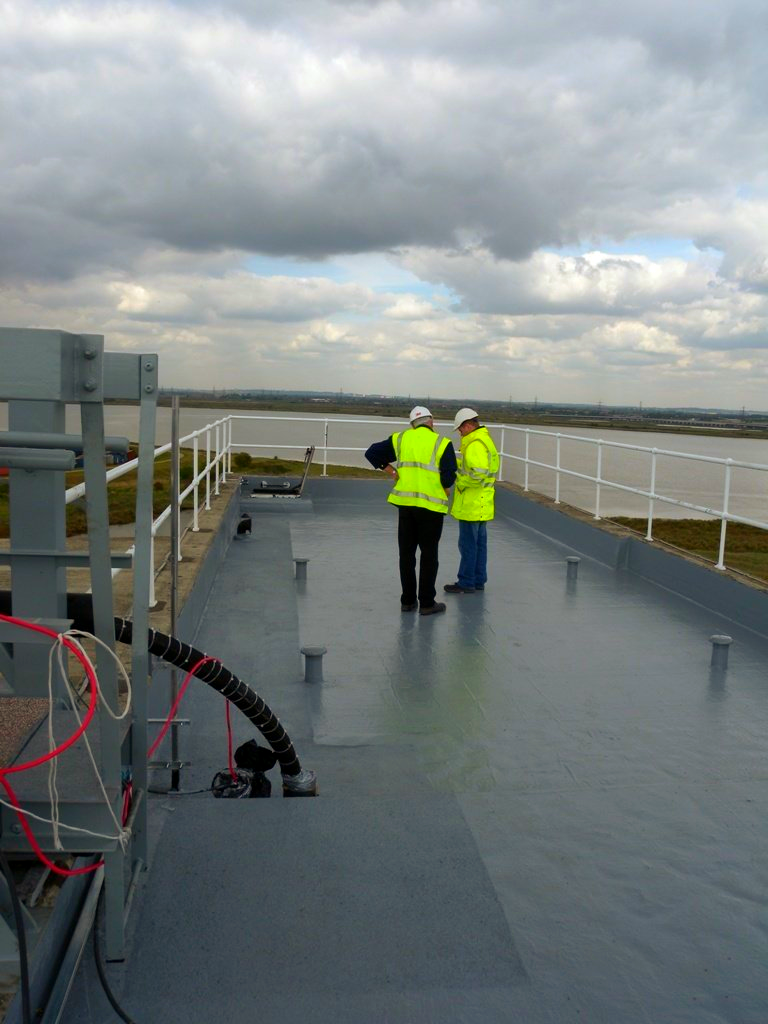Two men in yellow vests are standing on a roof overlooking a body of water.