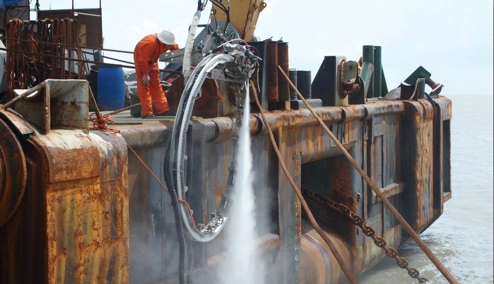 Man in orange suit operates machinery on rusty barge, spraying water.