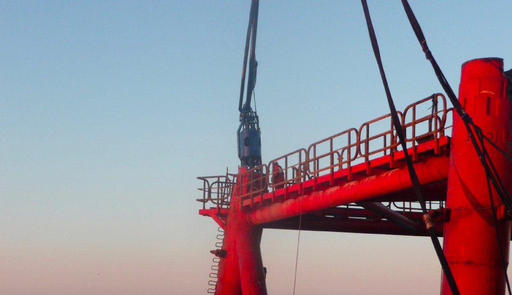 Red oil rig platform with equipment being lowered, against a blue and pink sky.