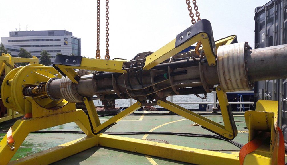 Yellow pipe-laying machine suspended by chains, holding a large pipe on a deck near a building.