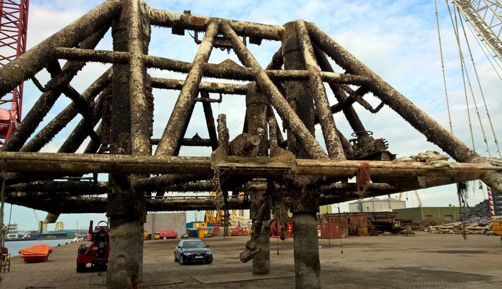 A large, barnacle-covered metal structure on concrete supports, in a harbor area, possibly being lifted by a crane.