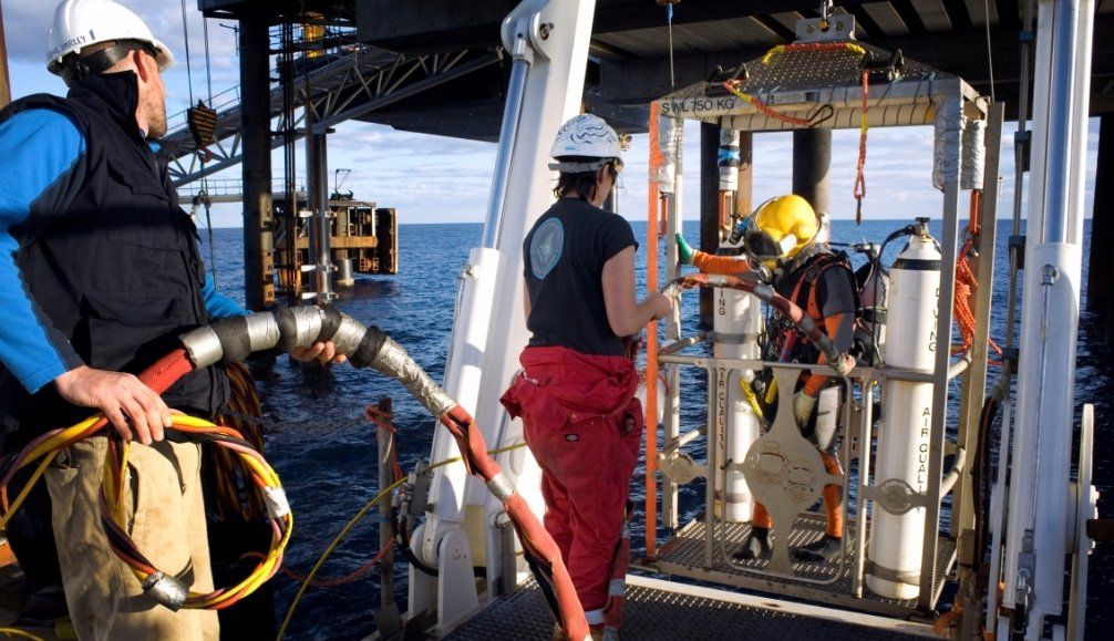 Workers assisting a diver in a cage; offshore oil rig in background.