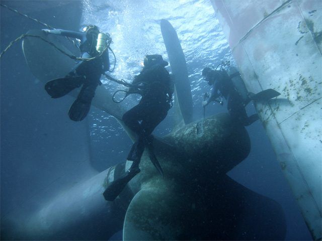 Three scuba divers near a large ship propeller underwater, inspecting or repairing it.