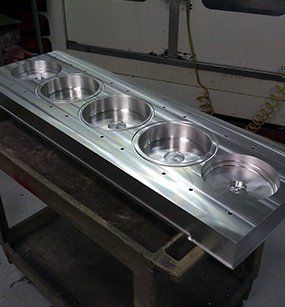 A long rectangular aluminum block with five circular machined cavities, resting on a workshop cart.