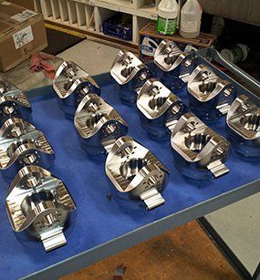 A tray on a work cart holds several shiny, machined metal components with cylindrical and bracket-like features.