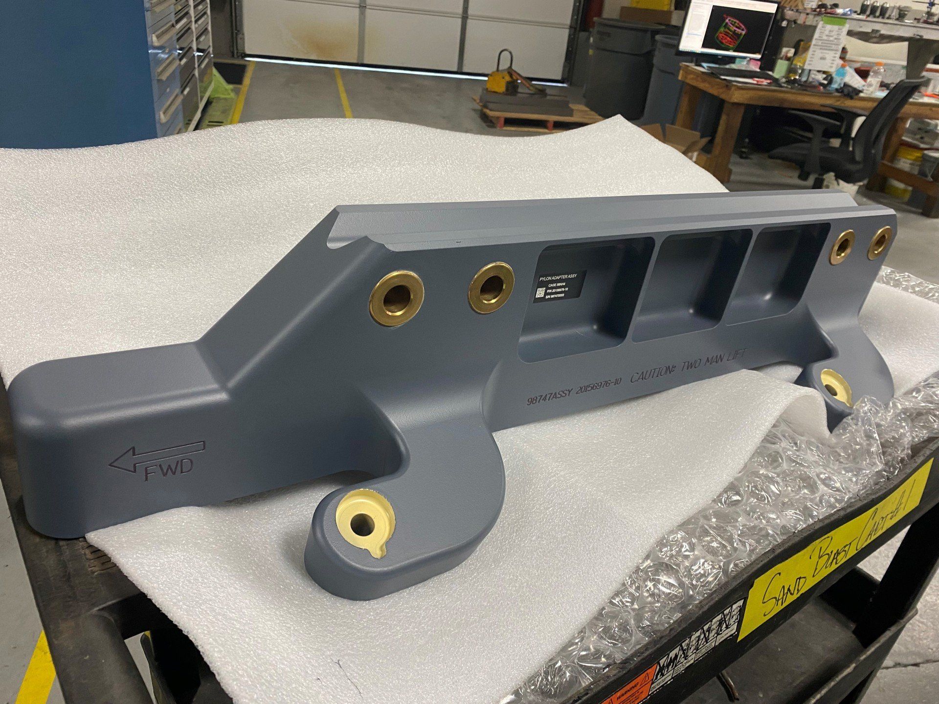 A gray, heavy-duty industrial metal bracket with brass-colored bushings, resting on protective foam in a workshop.