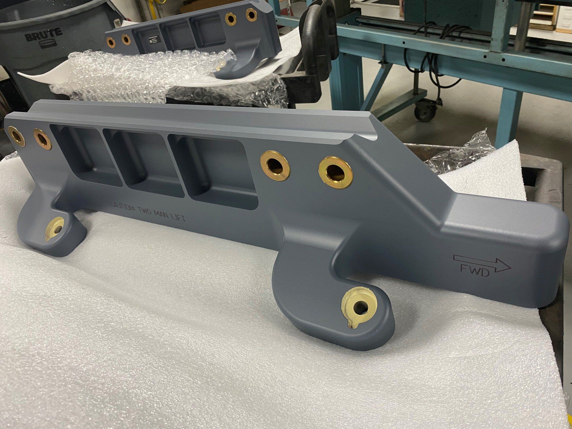 A grey, cast metal industrial machine component with brass-lined mounting holes, resting on foam packaging in a workshop.