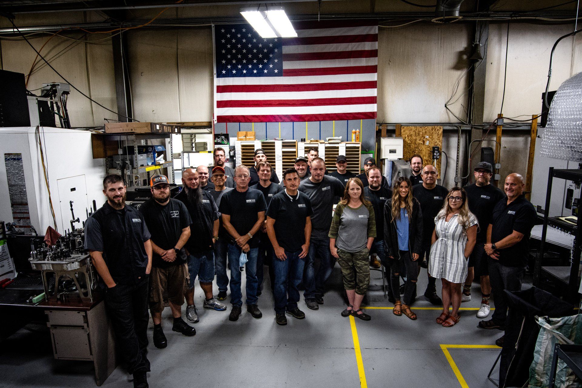 A group of employees stands together in a warehouse workshop beneath an American flag hanging on the back wall.