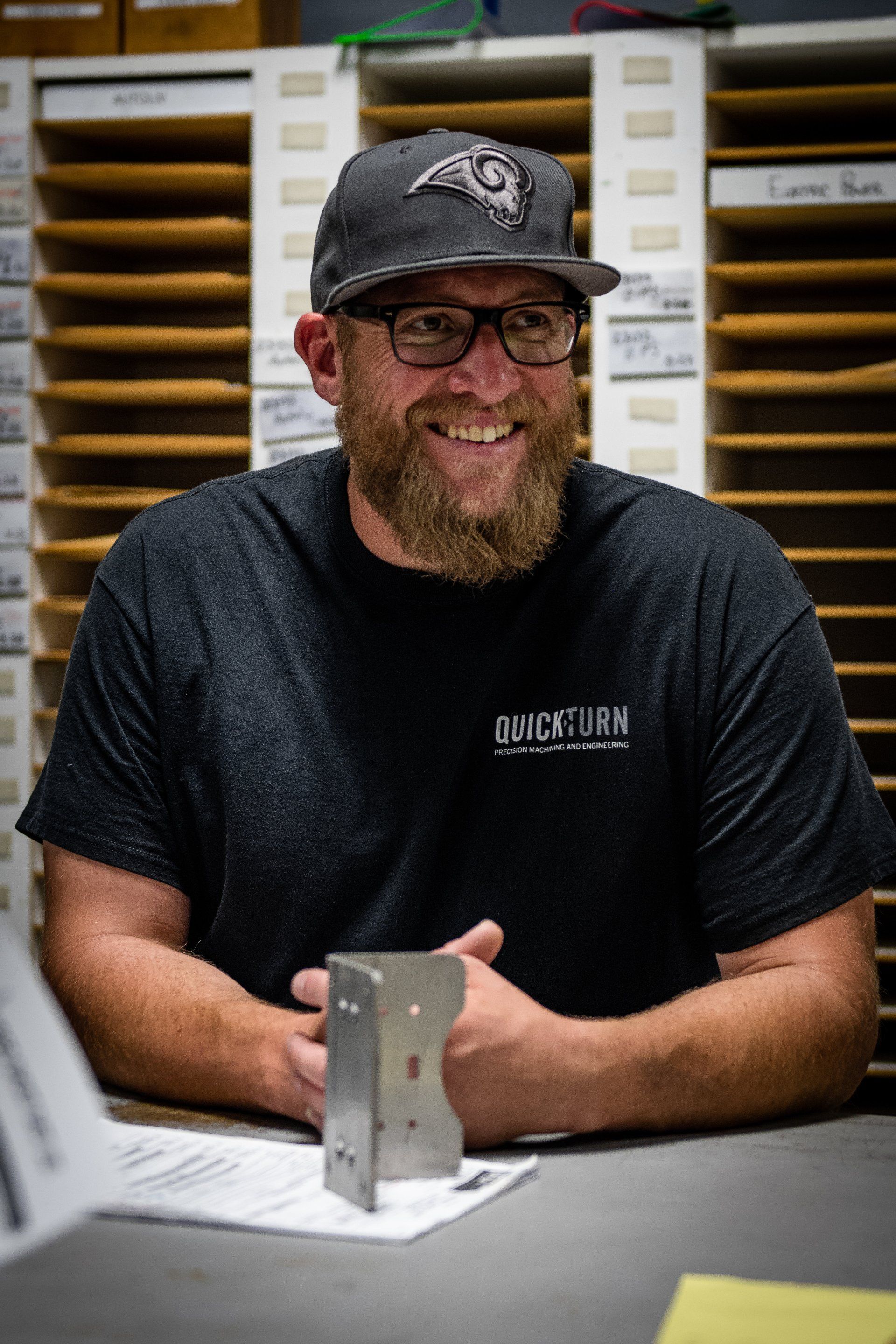 A smiling, bearded person in a baseball cap and dark t-shirt sits at a desk with a metal bracket in front of cubby shelves.