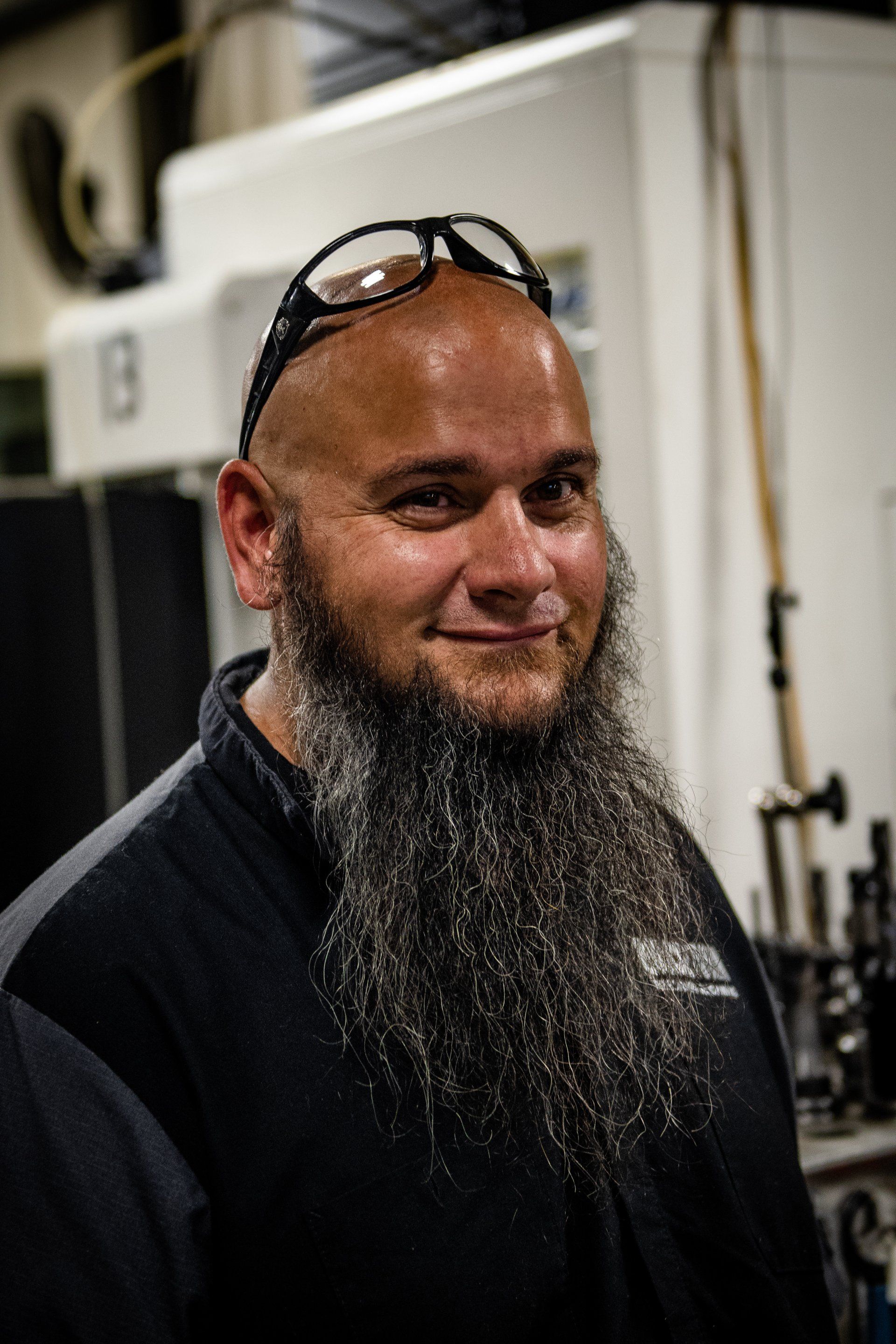 A bald person with a long, graying beard and safety glasses on their head smiles in a machine shop setting.