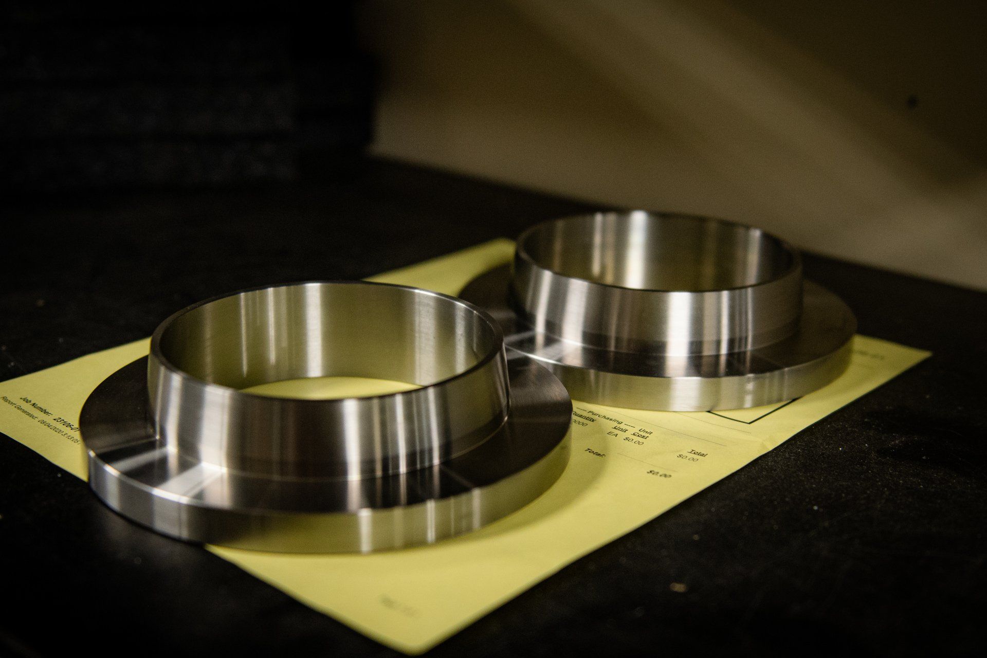 Two machined metal flanges with raised centers sit on a yellow piece of paper against a dark background.
