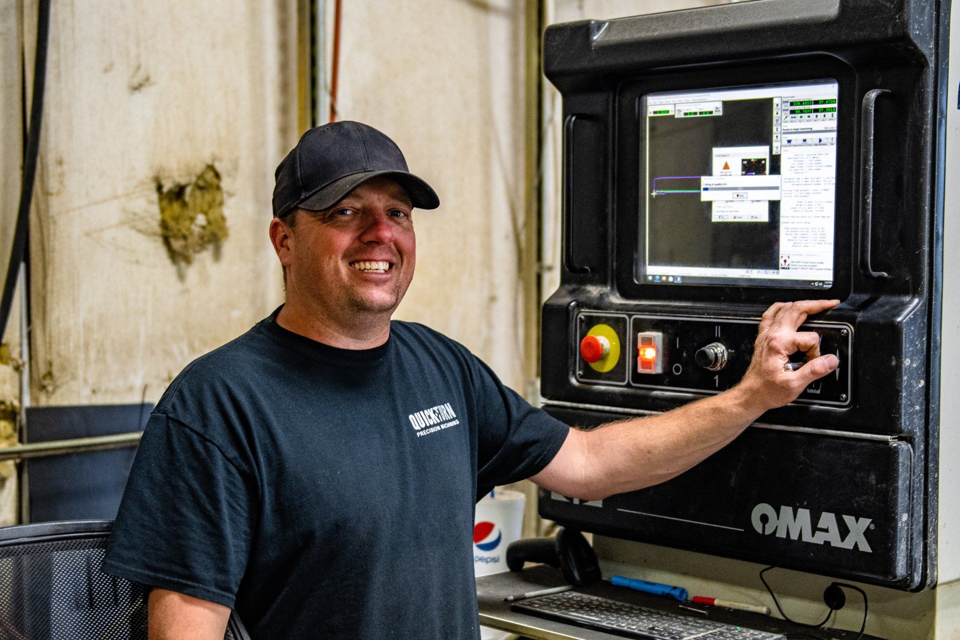 A person in a black cap and shirt smiles while operating an OMAX industrial machine control panel in a workshop.