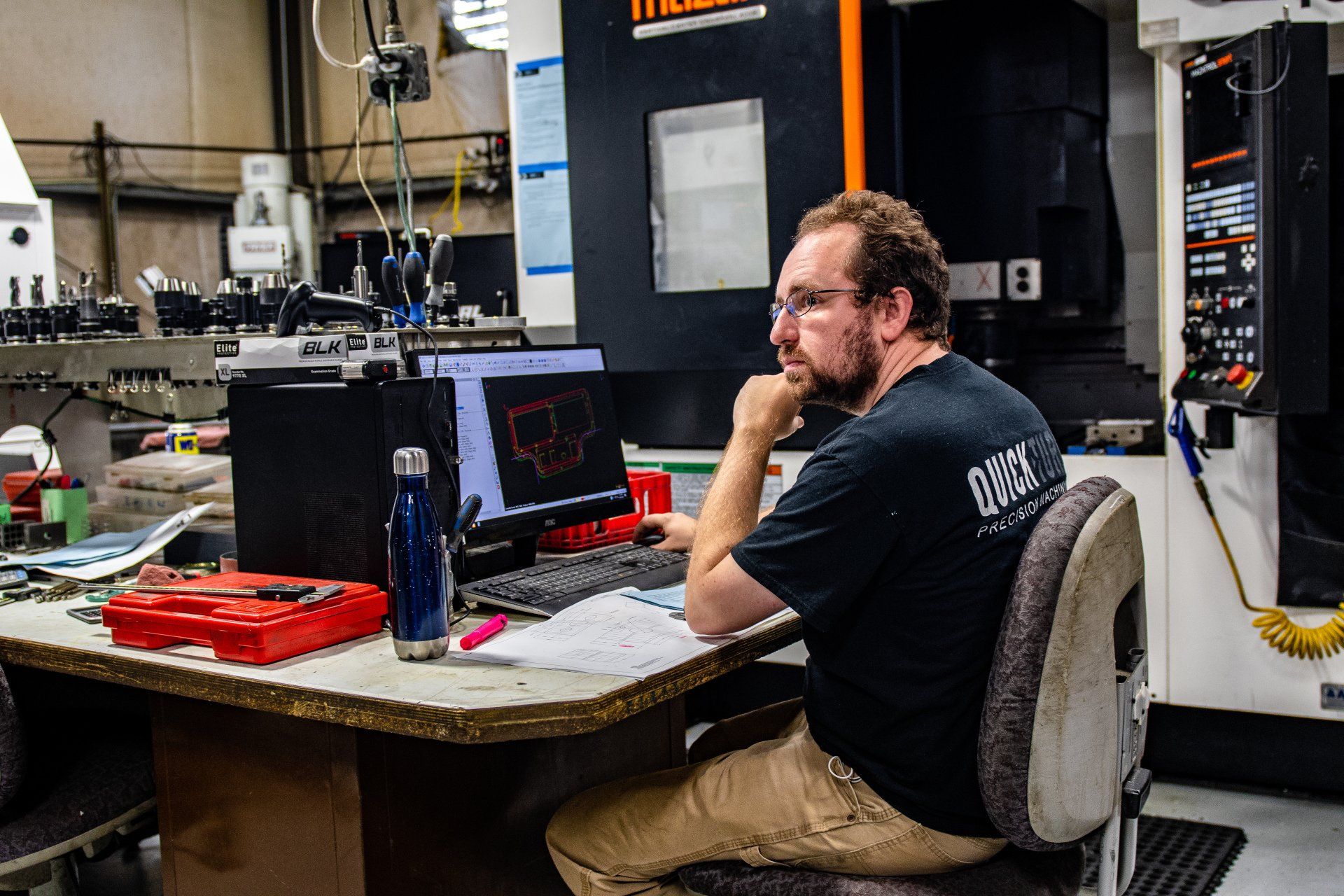 A person sitting at a computer workstation in a factory, reviewing a digital design on a screen near industrial machinery.
