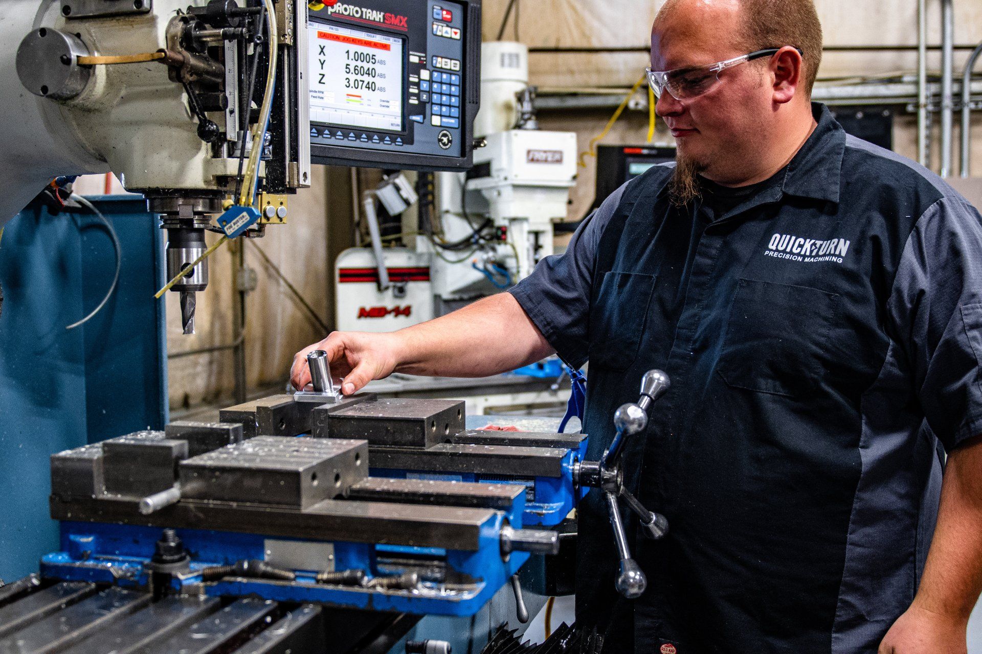 A technician wears safety glasses while adjusting a metal component held in a blue machine vise on a milling machine.