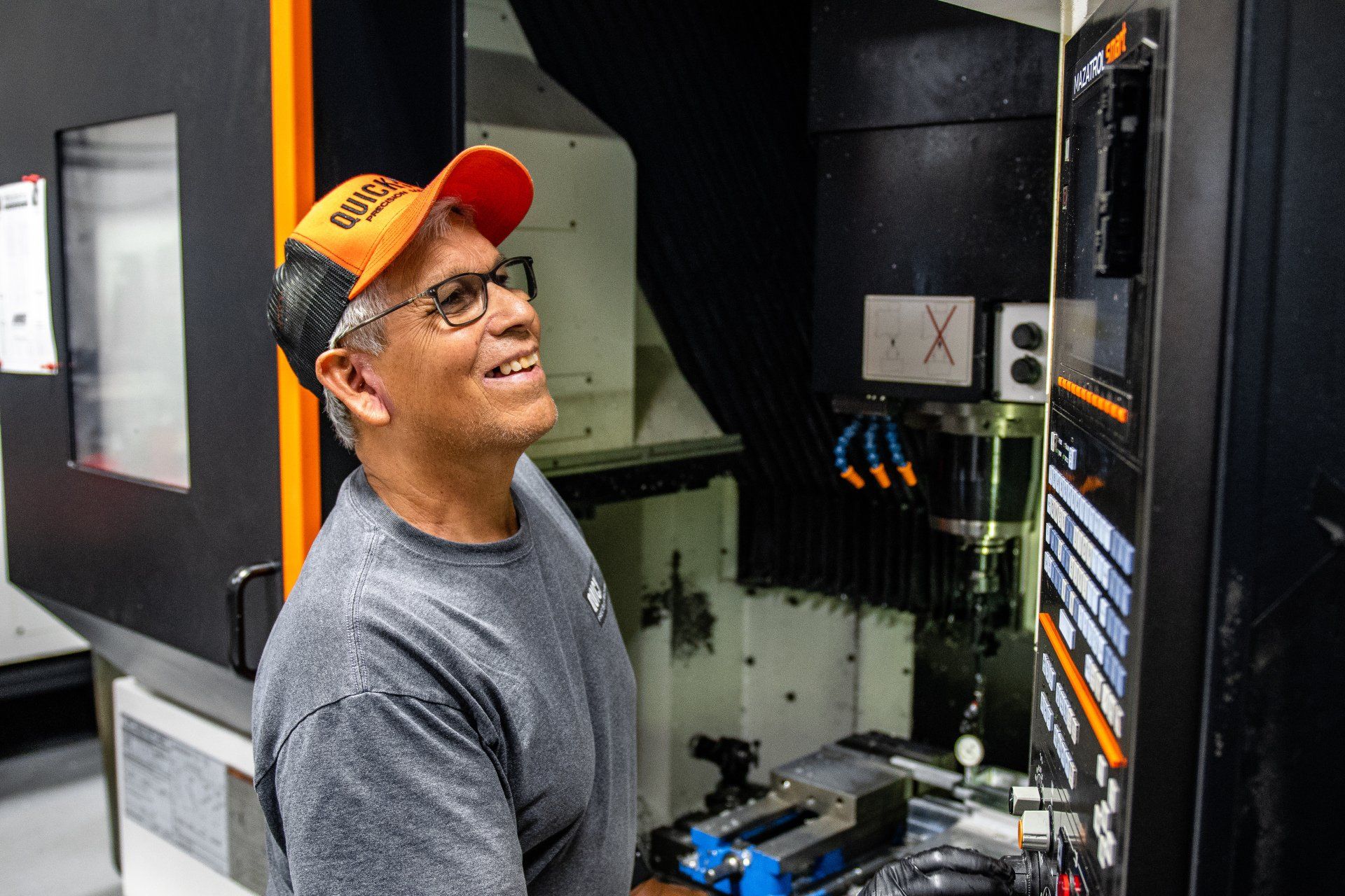 A smiling person wearing glasses and an orange cap operates a CNC machine in a workshop.