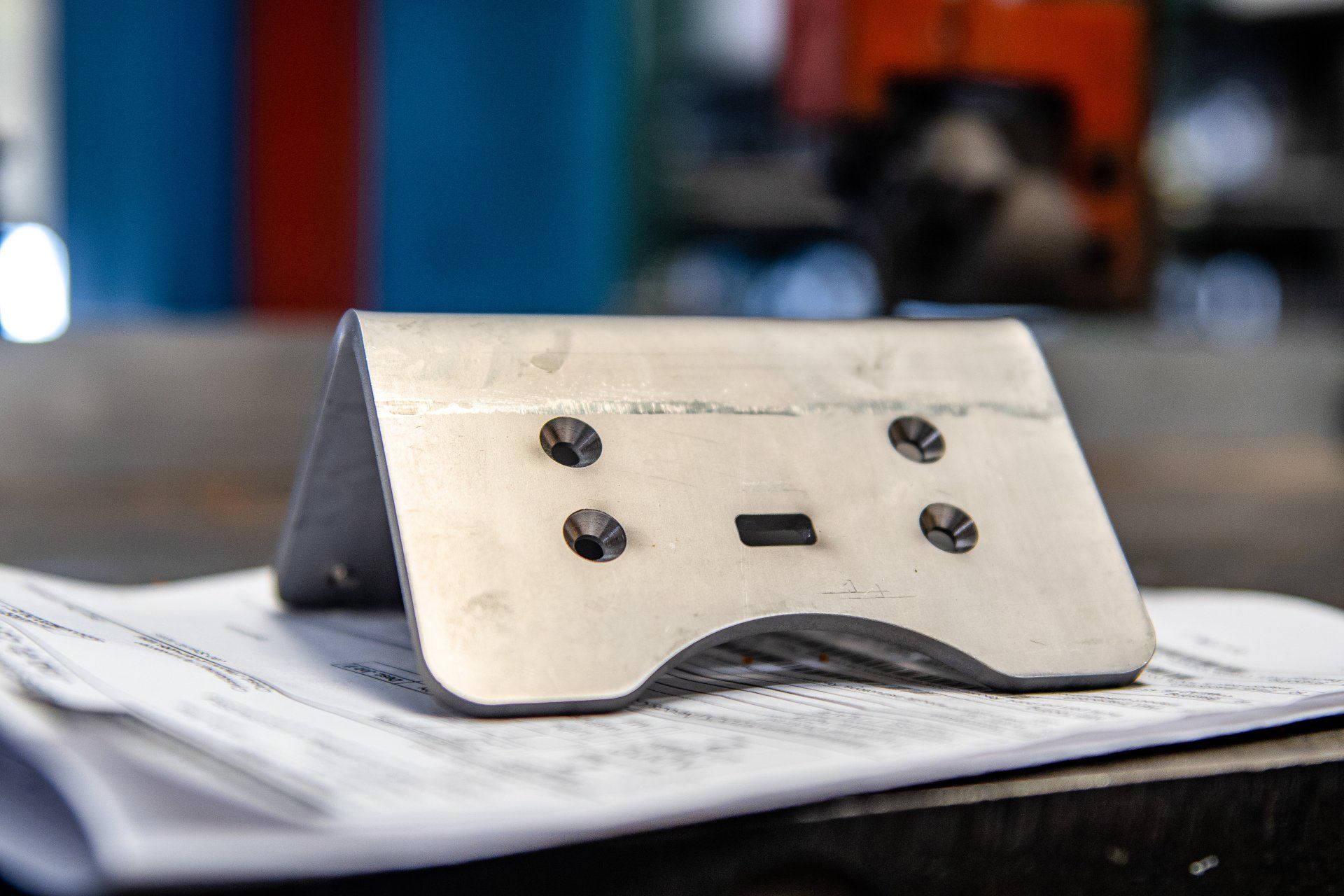 A bent metal bracket with four countersunk holes and a rectangular cutout, resting on engineering blueprints.