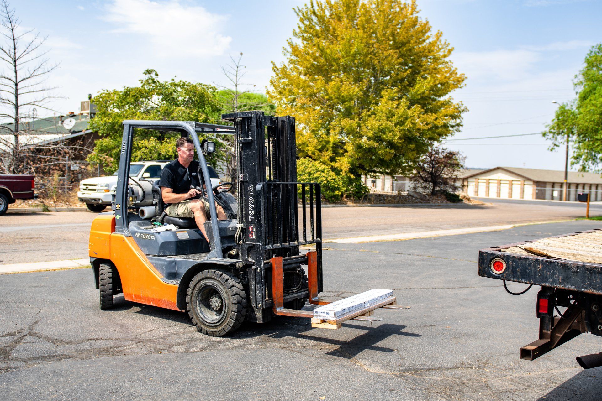 A person driving an orange forklift, carrying a pallet toward the back of a flatbed truck on an outdoor paved lot.