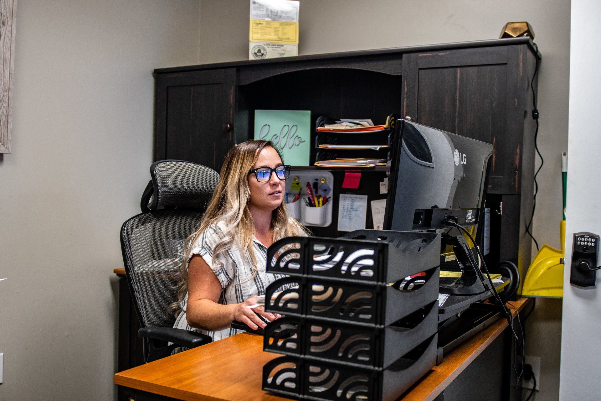 A person sits at an office desk using a computer, with a stack of desk trays in the foreground.