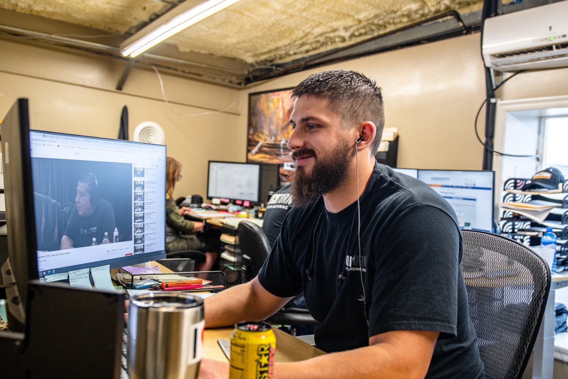 A bearded man with a headset smiles while working at a computer desk in an office setting.
