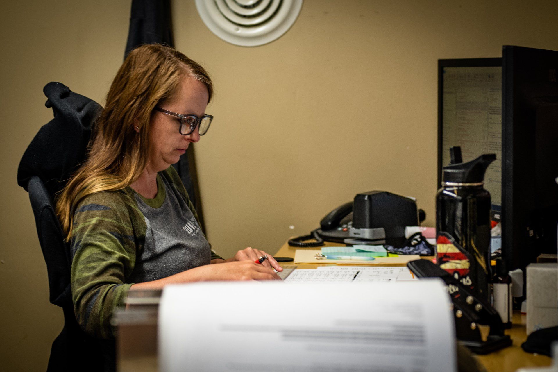 A person wearing glasses and a long-sleeved shirt sits at an office desk working on documents near a computer monitor.