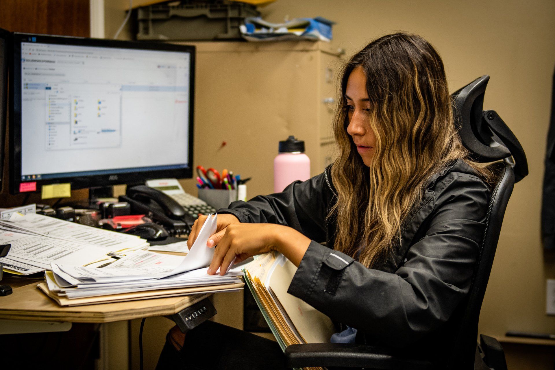 A person in a black jacket sits at a desk, reviewing documents and a binder in front of a computer monitor in an office.