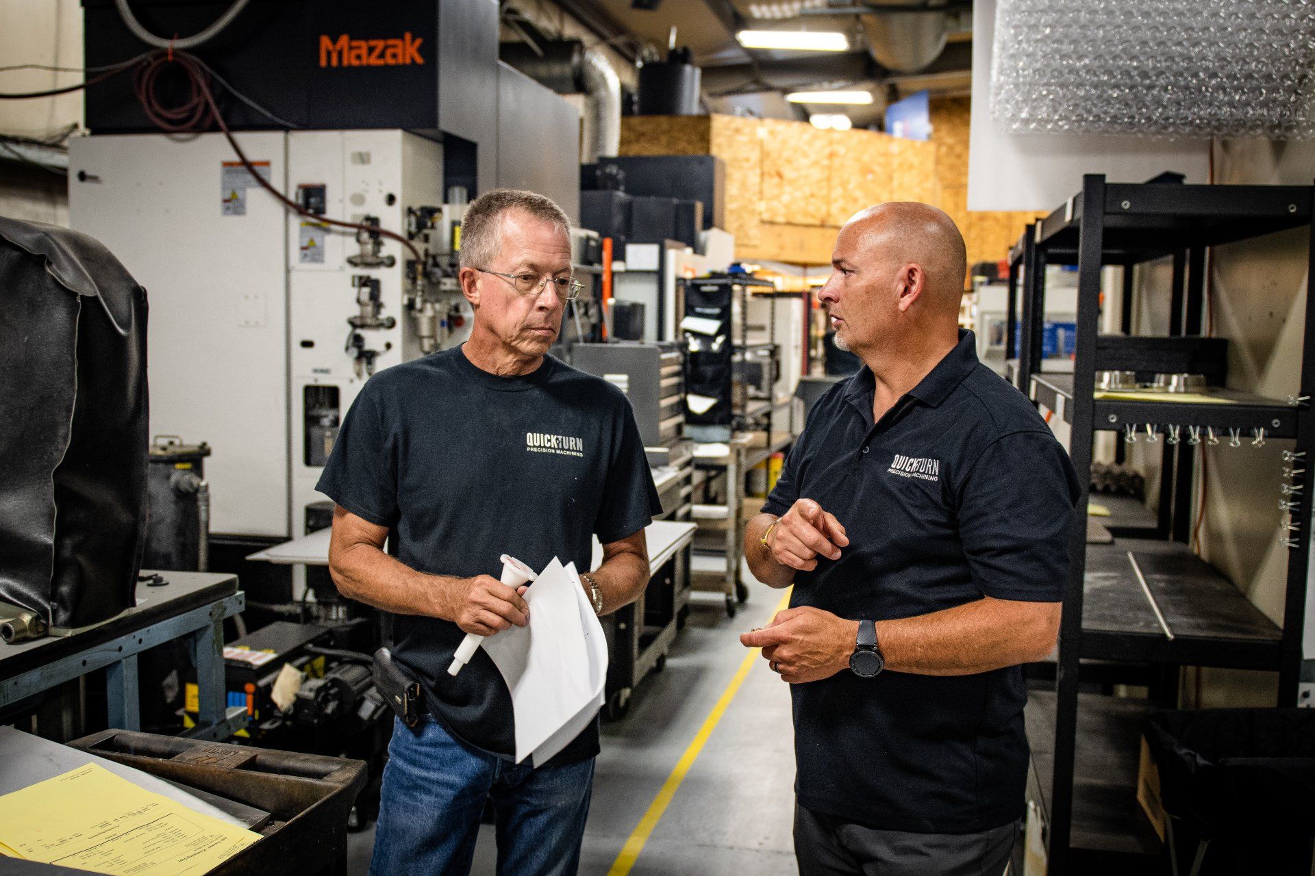 Two professionals in dark shirts discuss documents while standing in a manufacturing shop with heavy machinery.