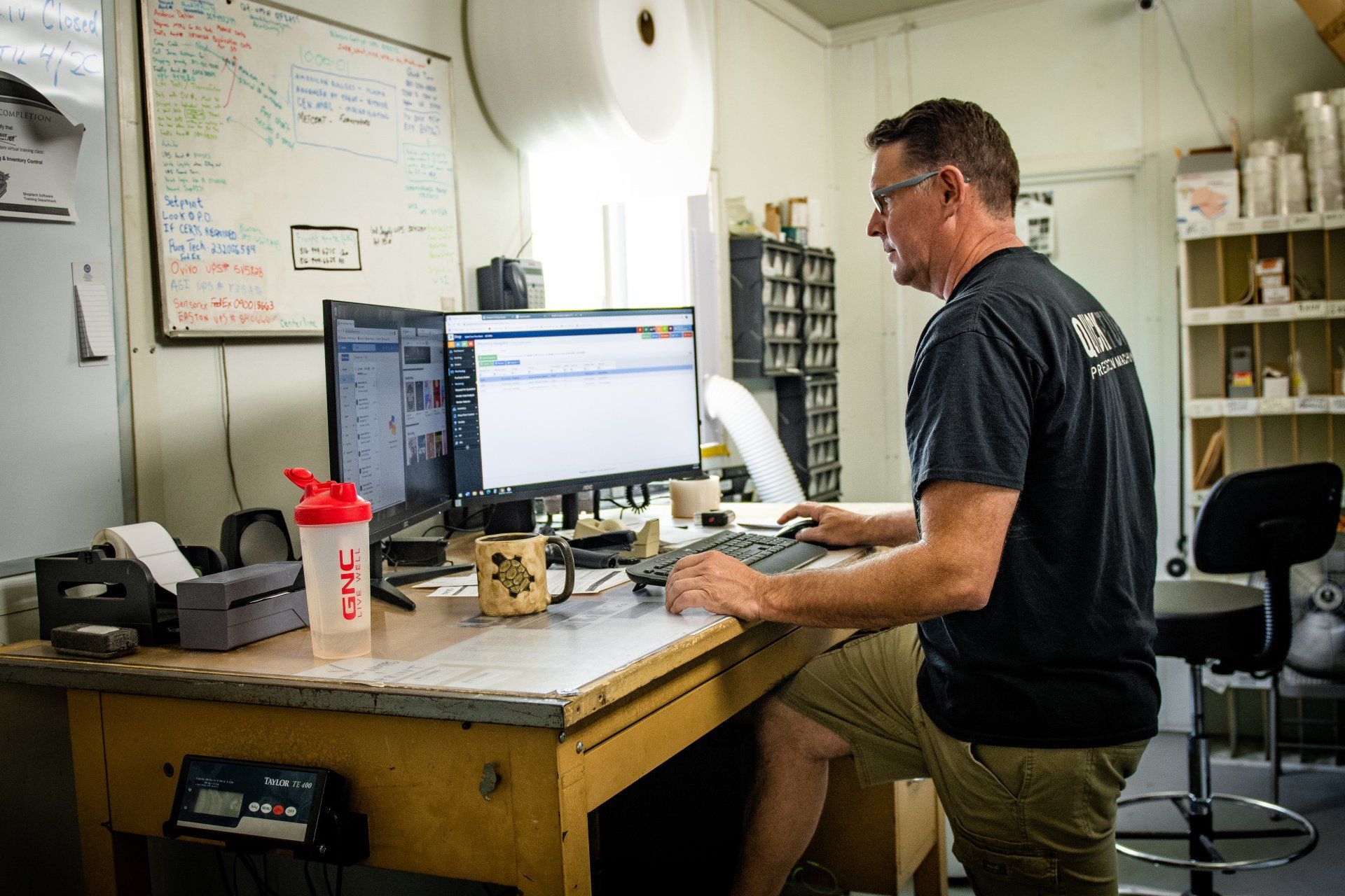 A person in a black t-shirt works at a dual-monitor computer setup in an office or warehouse setting.