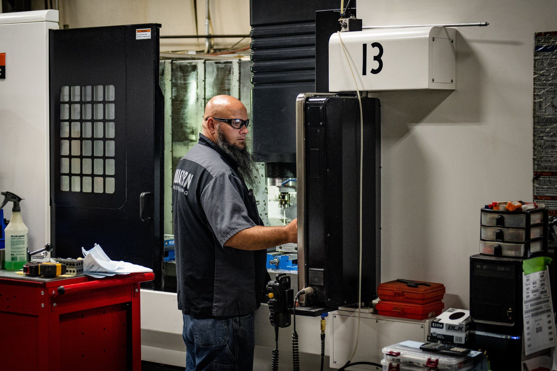A person wearing safety glasses and a gray uniform operates machinery at a workstation marked with the number 13.
