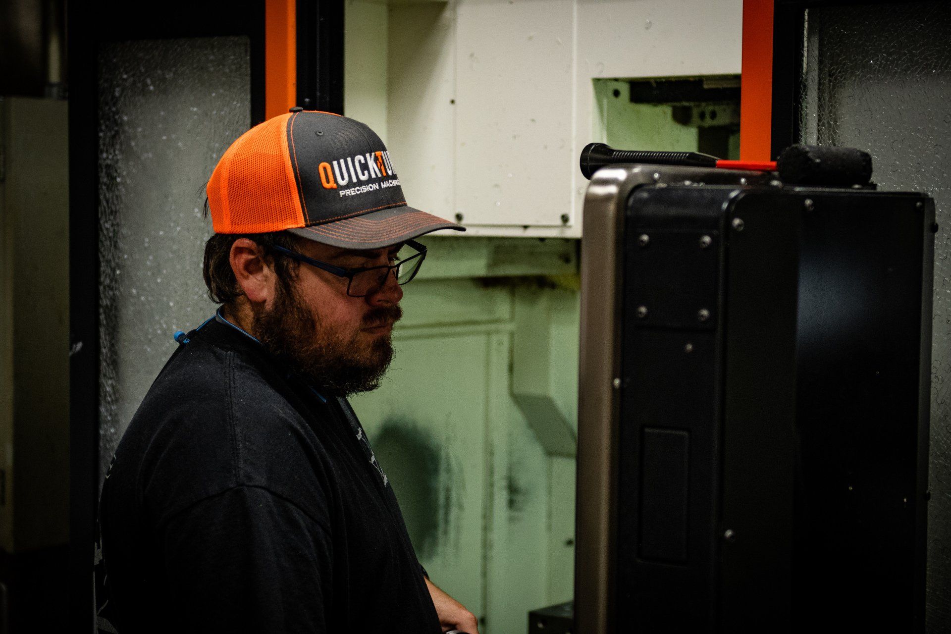 A bearded person wearing an orange and gray baseball cap operates industrial machinery in a workshop.