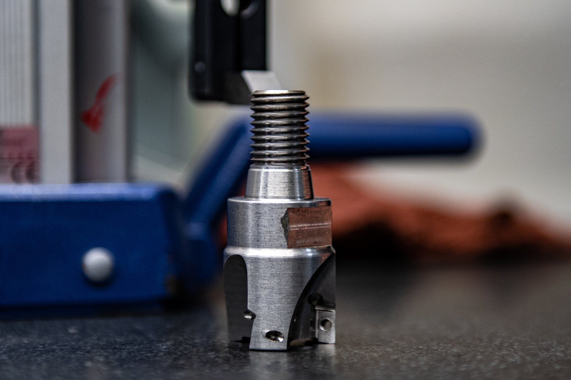 A metal milling cutter tool with a threaded shank stands vertically on a dark surface in a workshop setting.