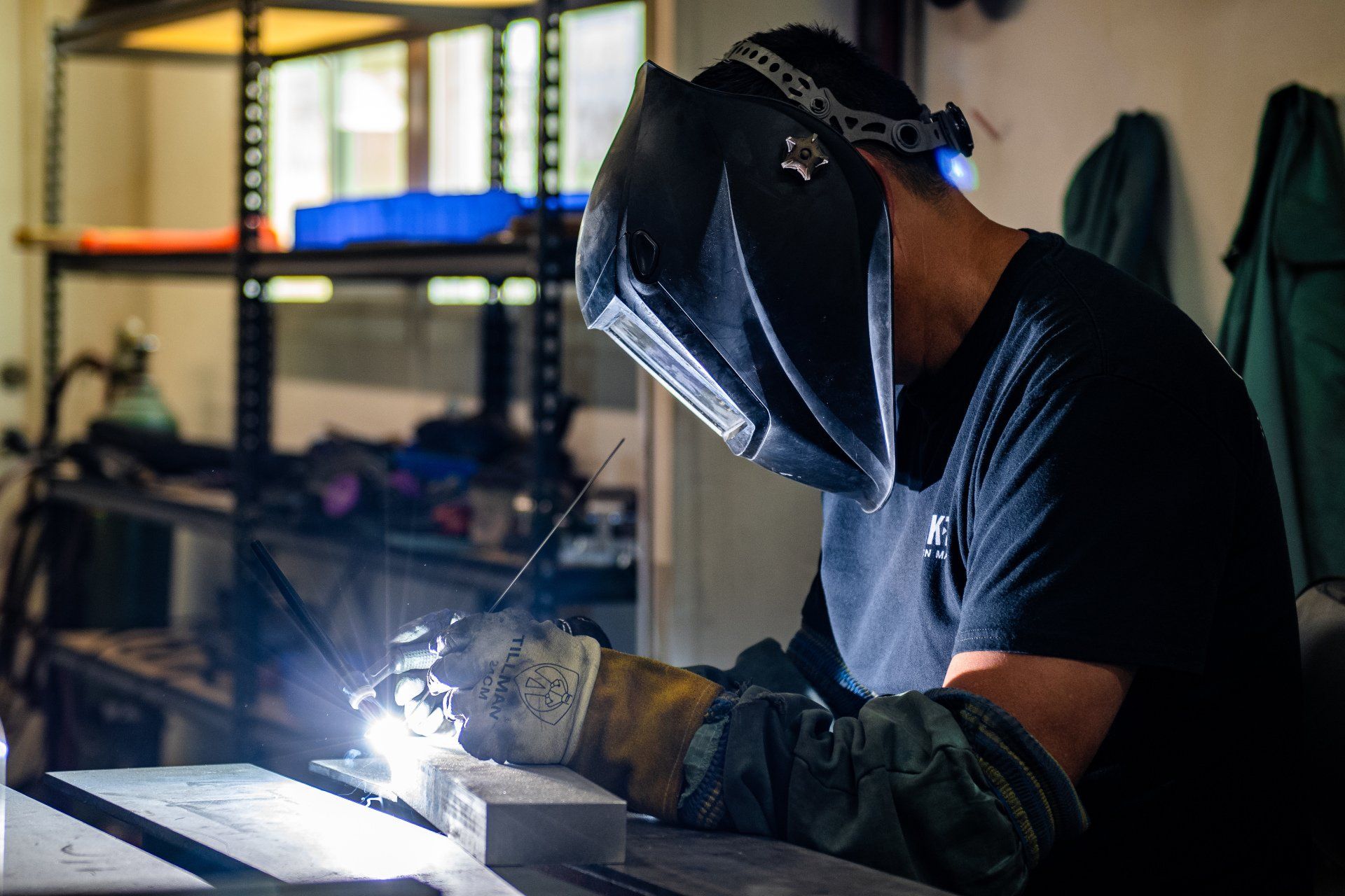 A person wearing a protective helmet and gloves arc-welding metal in a workshop.