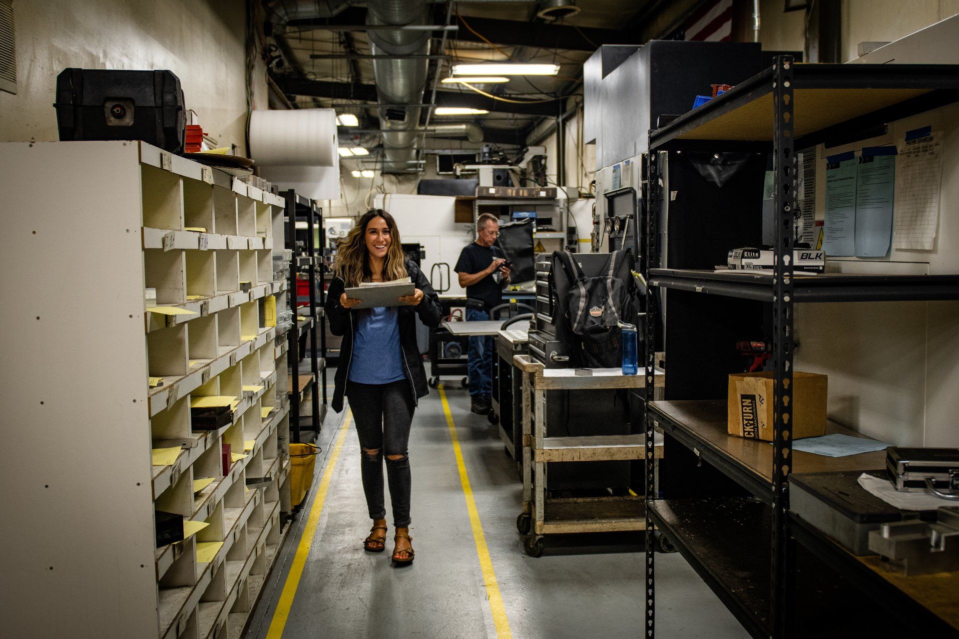 A person smiling while carrying a box through an industrial storage area with metal shelves and yellow floor markings.