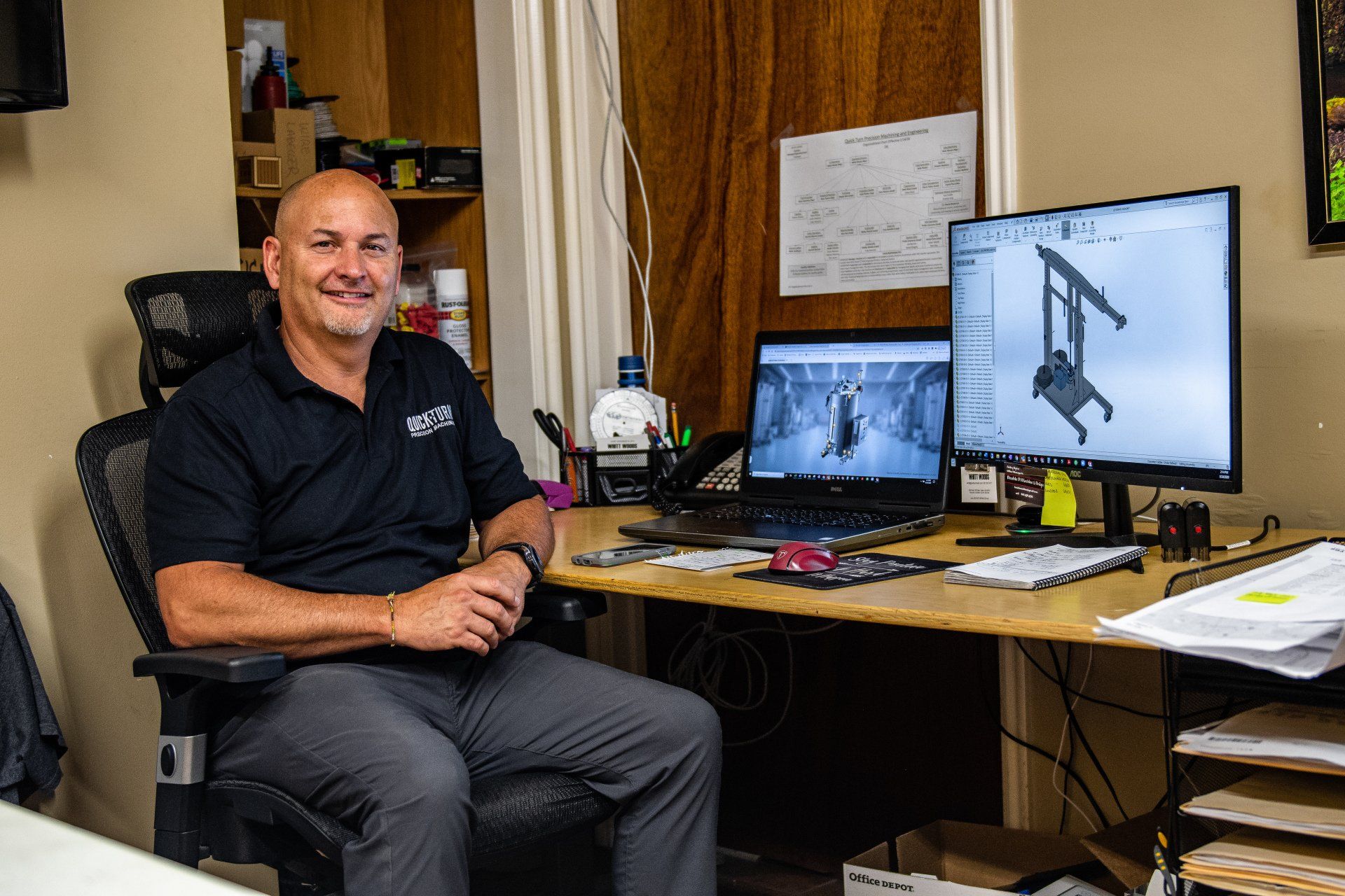 A person sitting in an office chair in front of a desk with two computer screens displaying engineering designs.