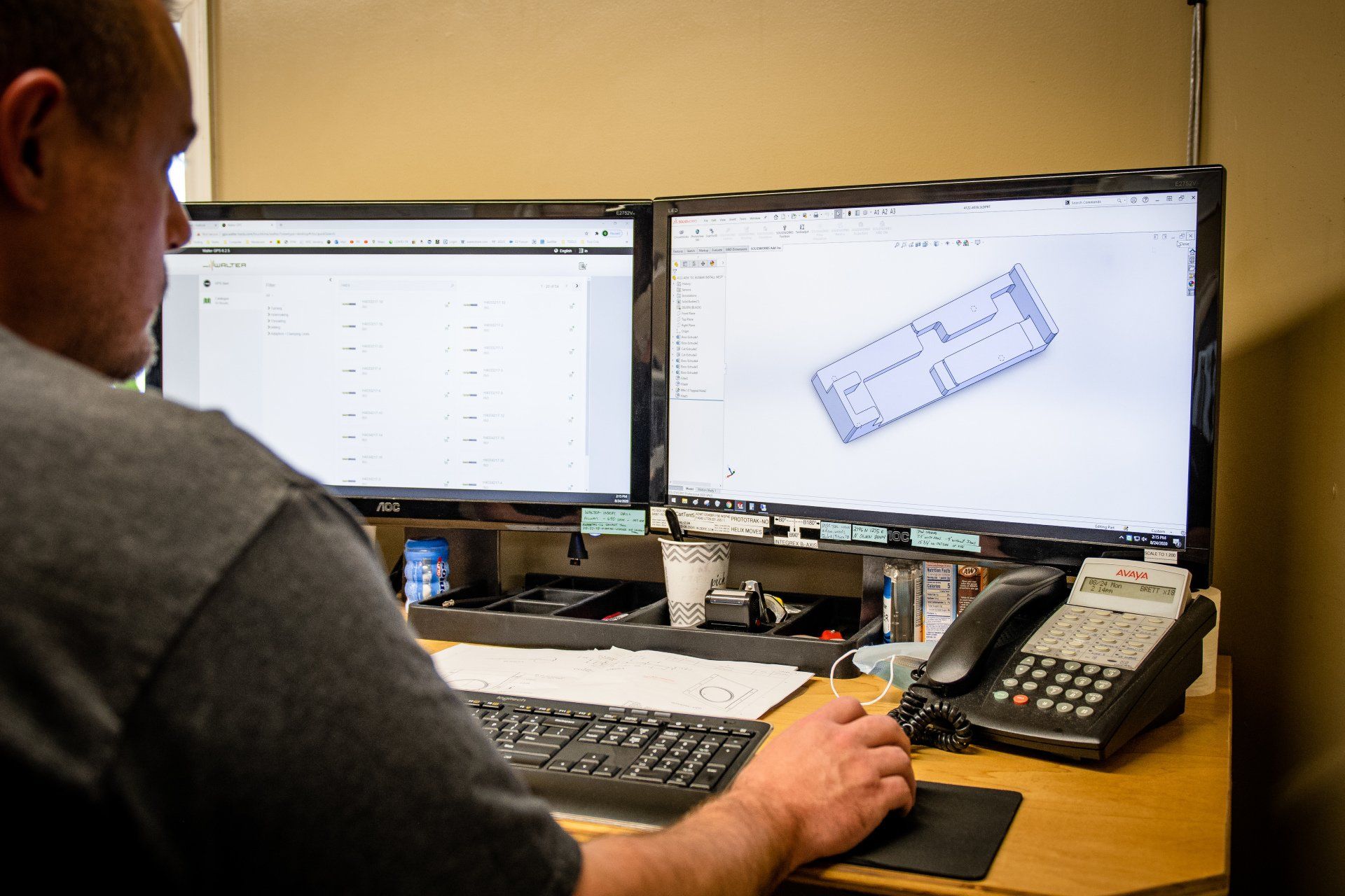 A person sits at a desk with two monitors displaying technical floor plans and data, using a computer mouse and phone.