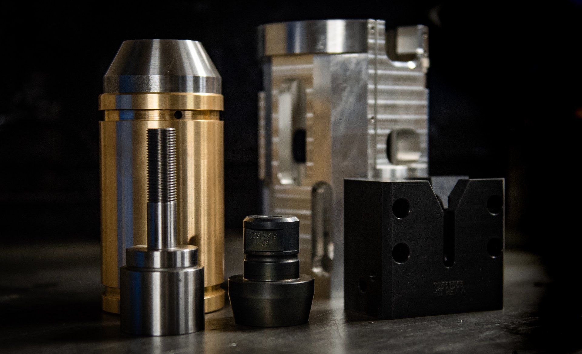 Precision machined metal components, including brass, steel, and black hardware, arranged on a dark workshop surface.
