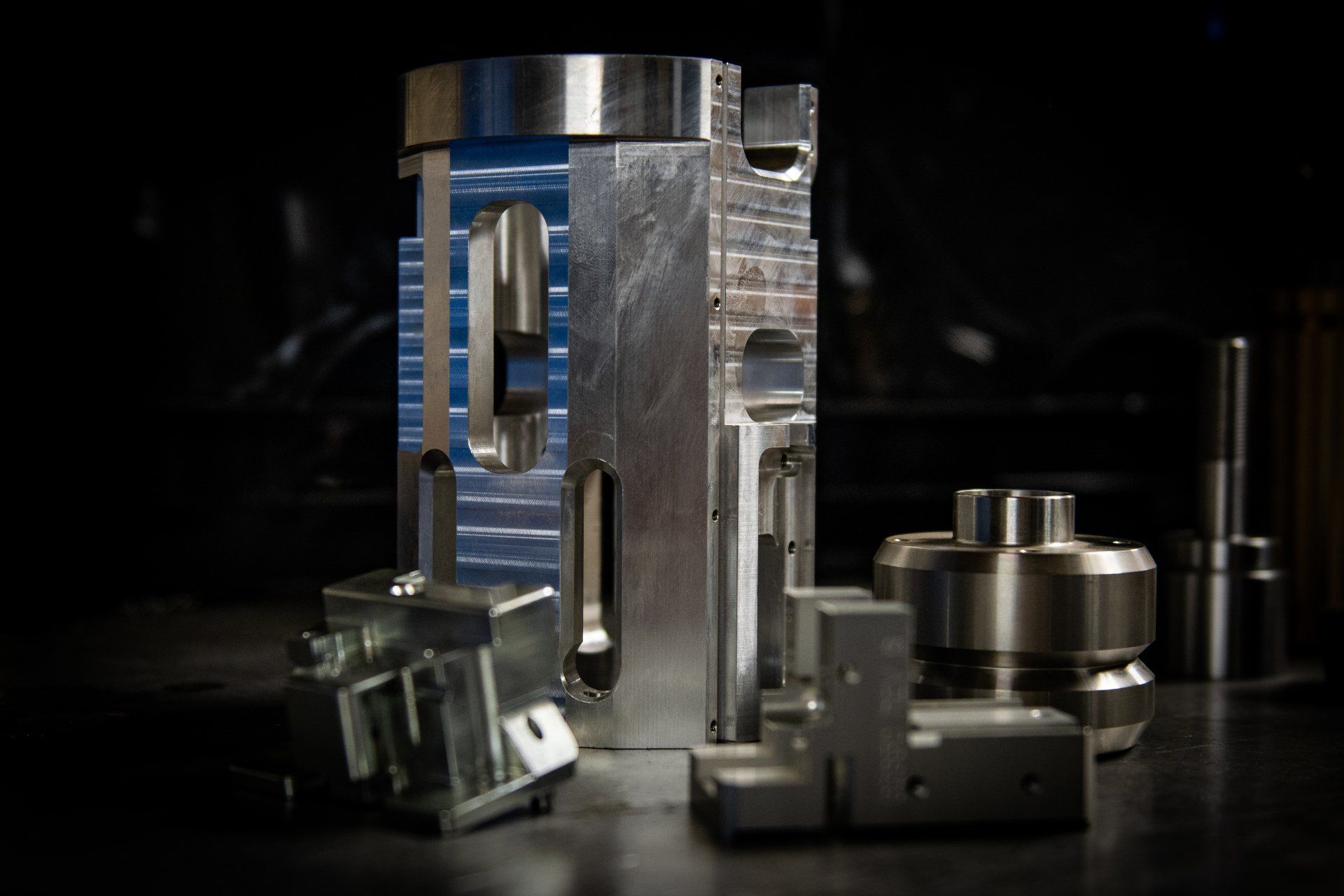 Several precision-machined metal components sit on a dark surface, including a tall, slotted cylinder and small parts.