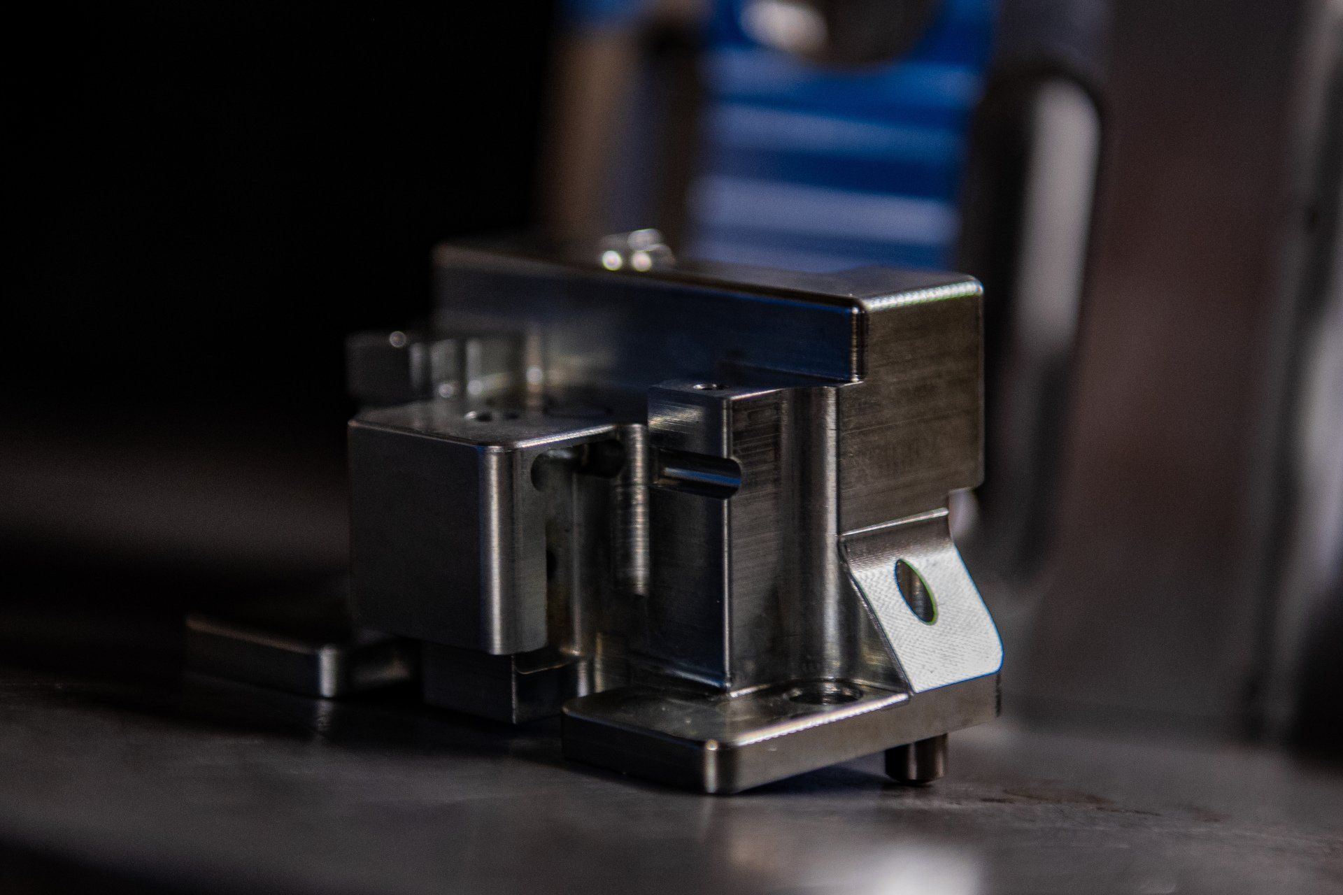 A metallic, machined mechanical component with angled surfaces and a mounting hole, resting on a dark surface.