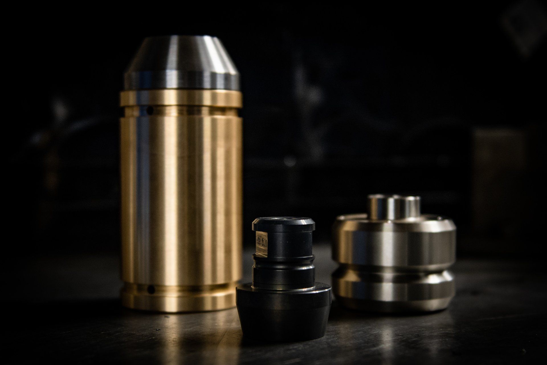 Three metal machined components—one large brass cylinder and two smaller, distinct metallic parts—on a dark surface.