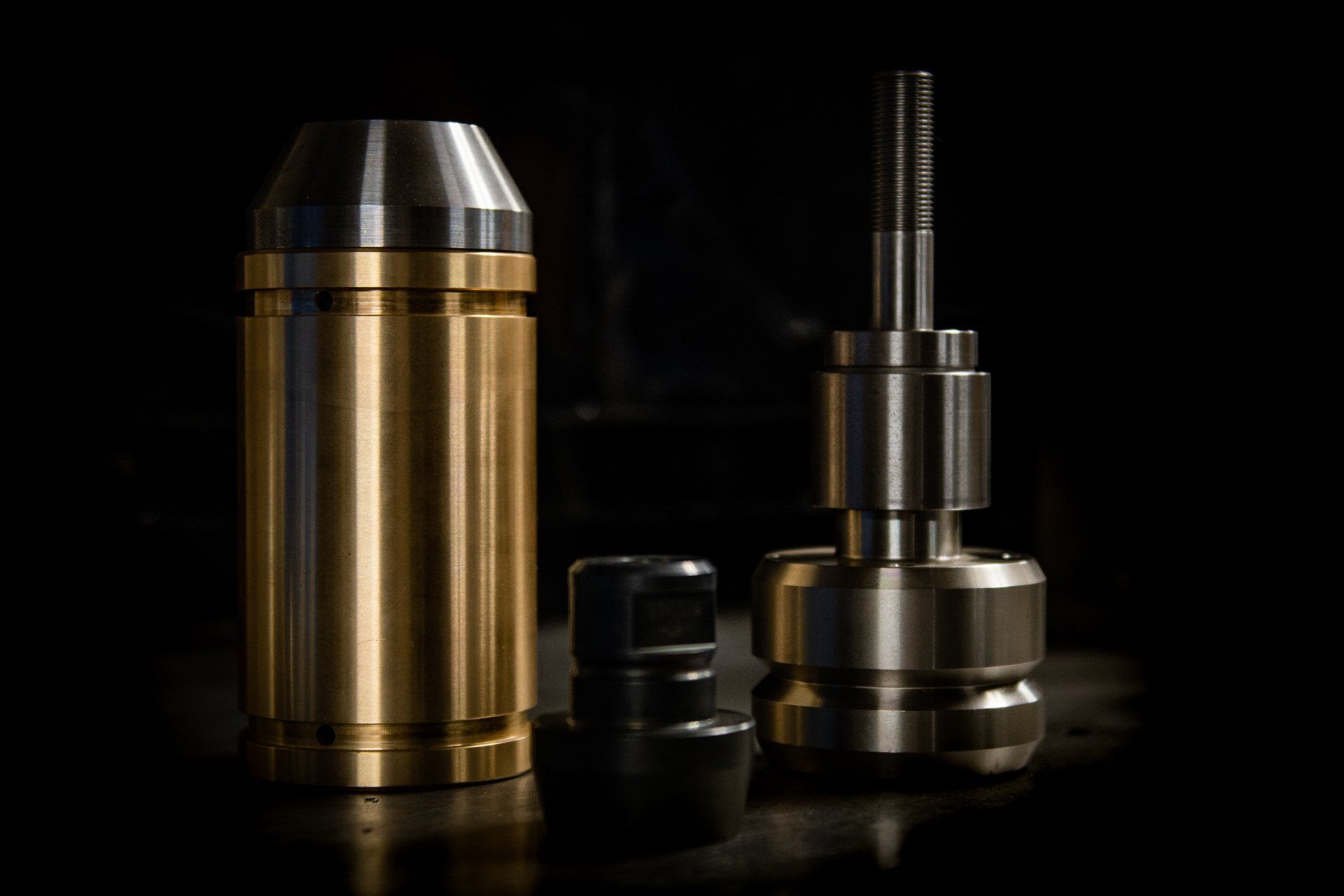 Three industrial metal components, including a brass cylinder and two steel parts, sit on a dark surface.