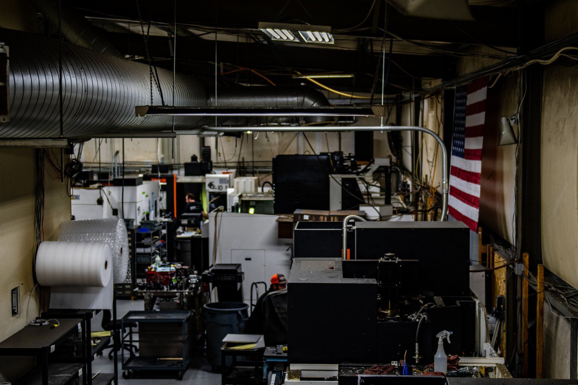 Industrial workshop interior with machinery, large rolls of plastic wrap, and an American flag hanging on the wall.