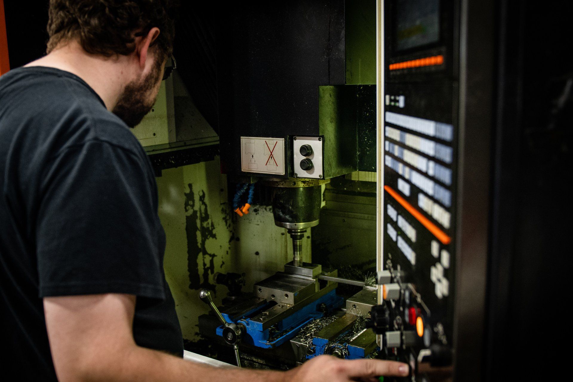 A person in a black t-shirt operates the control panel of a CNC milling machine in a workshop.