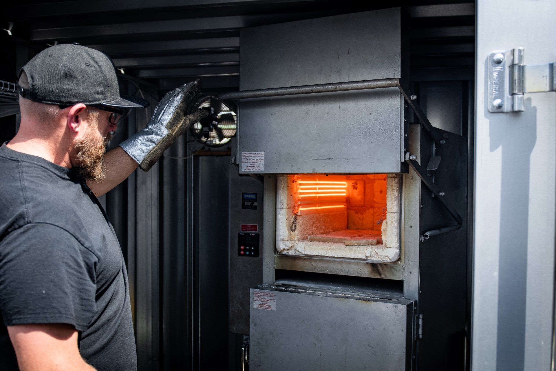A person wearing a heat-resistant glove opens the door of an industrial furnace, revealing glowing red-hot elements.