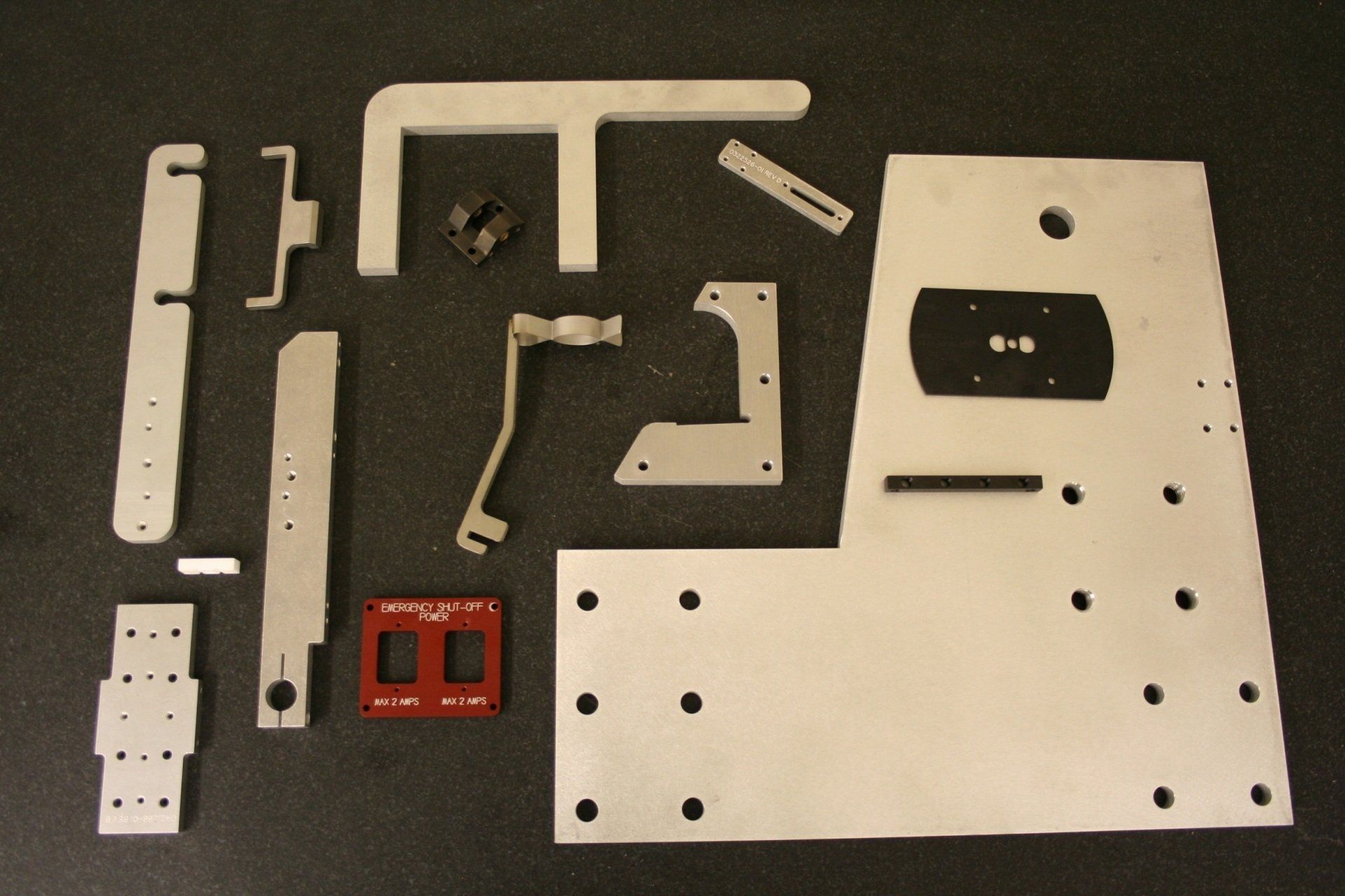 A collection of assorted metal and plastic mechanical parts, including plates, brackets, and a red square piece, on a mat.