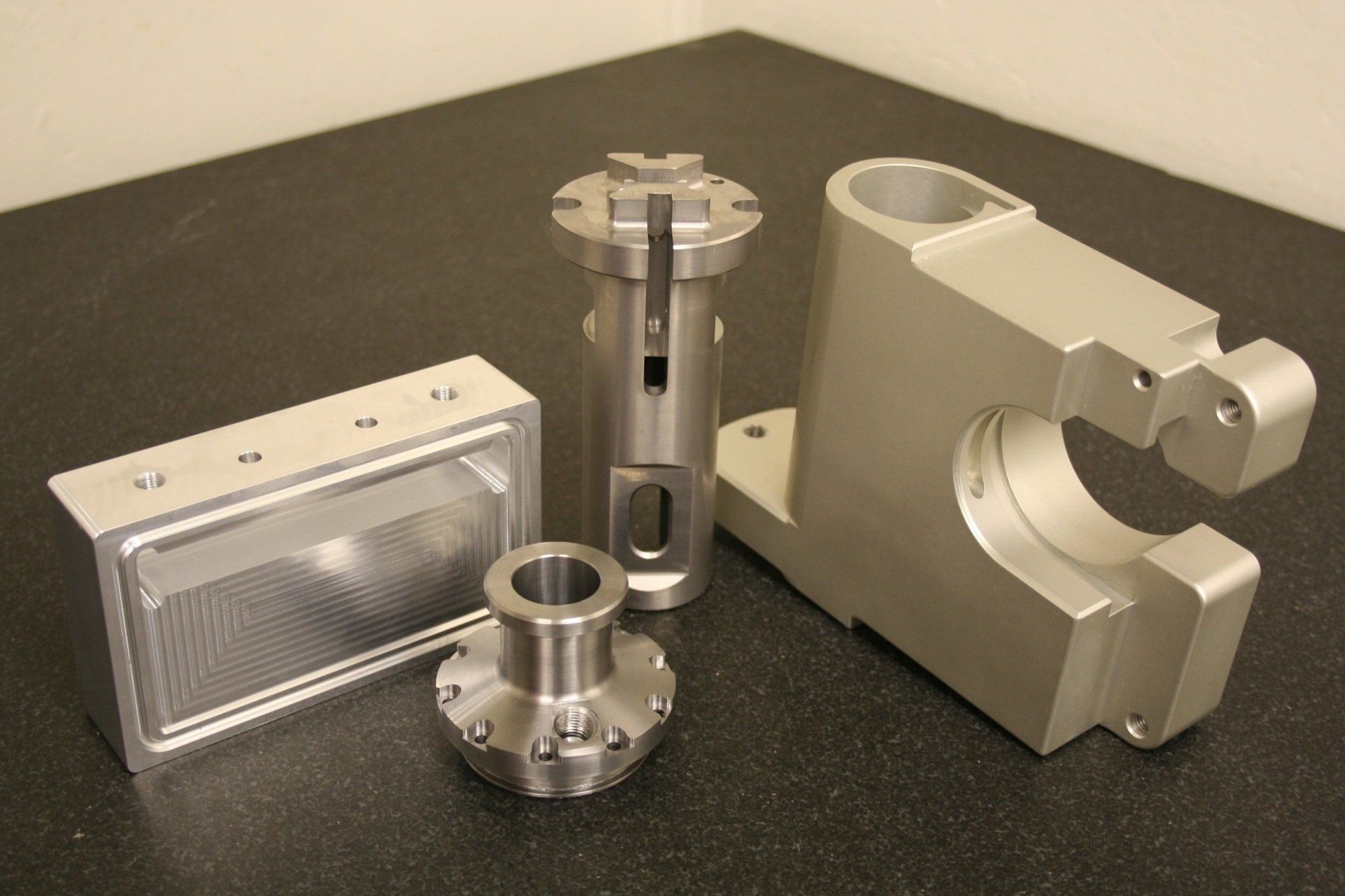 Four machined metal parts of varying shapes and sizes arranged on a dark, textured surface.