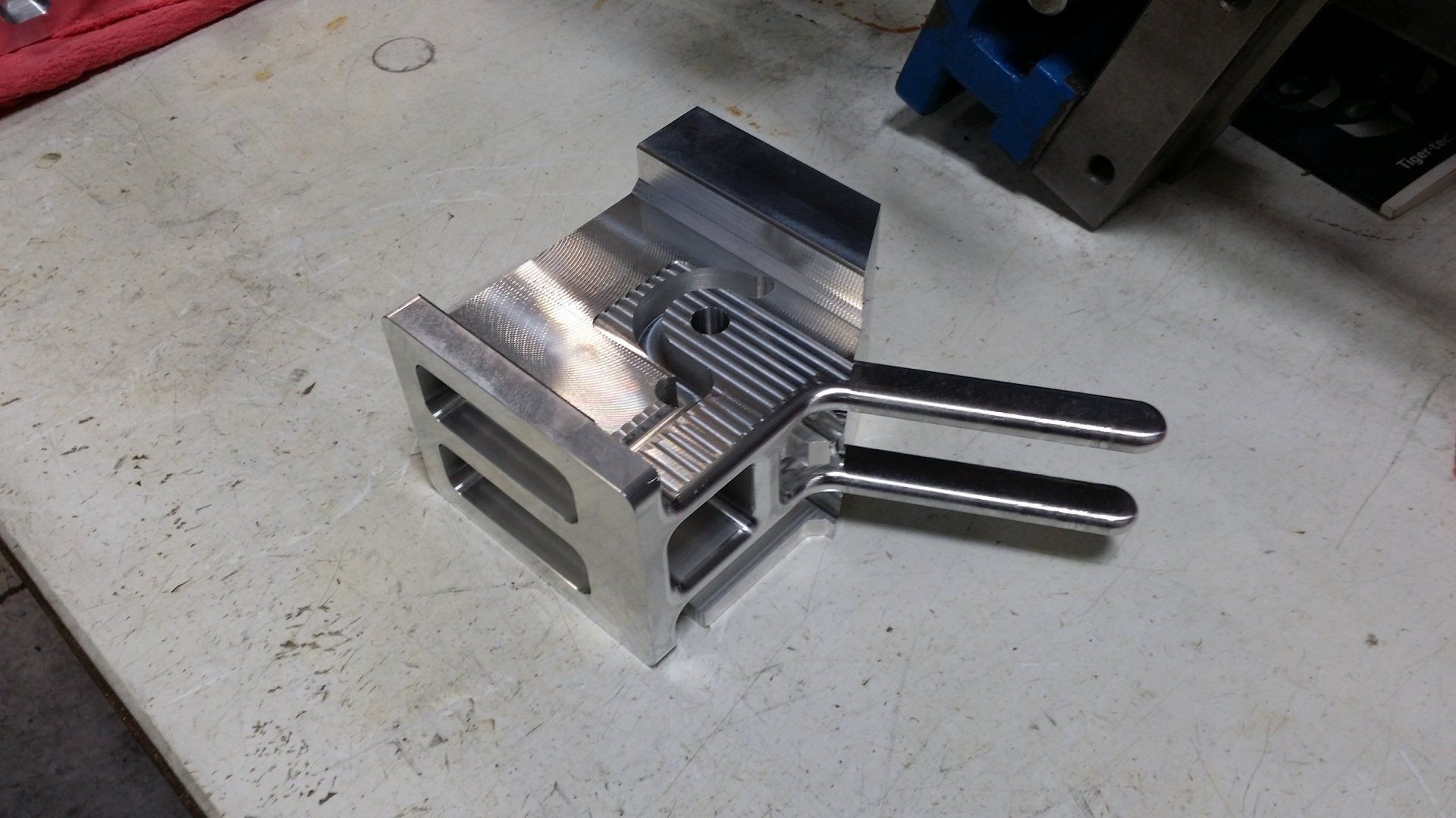 A machined aluminum part with two parallel metal arms protruding from one side, resting on a workshop workbench.