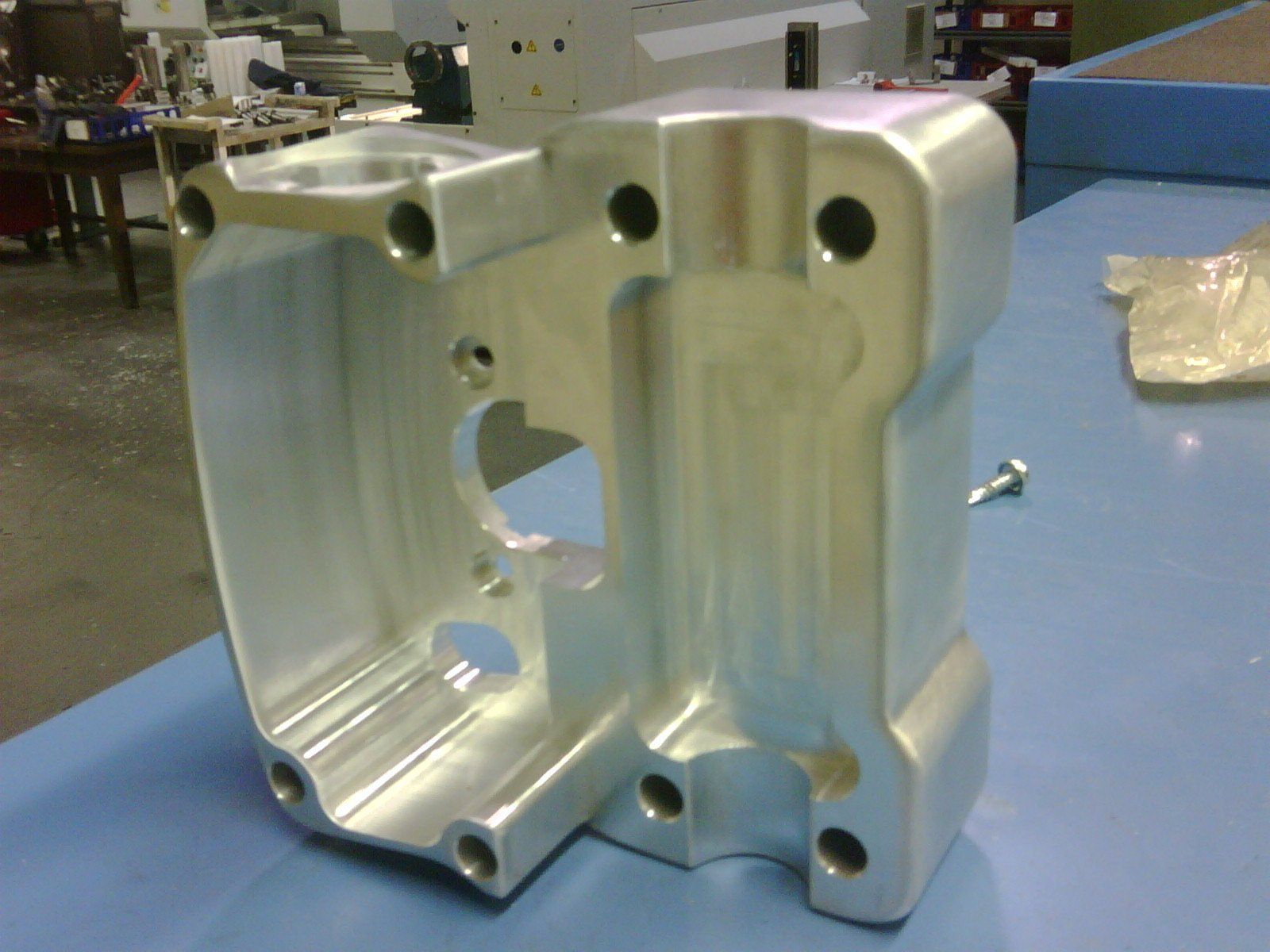 A machined aluminum mechanical component with multiple bolt holes, resting on a blue surface in a workshop setting.